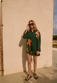 Groen overhemd met bloemenprint, met lange mouwen en een comfortabele pasvorm. G draagt het met zwarte slippers tegen een gestructureerde beige muur.