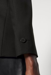 Manche de veste noire sur mesure en tissu texturé. Un bouton rond noir avec le logo "HUGO" est visible au niveau du poignet.