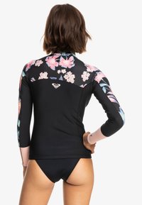 Tajón negro con estampado floral en los hombros y las mangas. Cuenta con mangas largas, un respaldo sin cremallera y un logo en la parte posterior del escote.