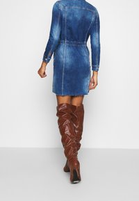 Denimklänning i blått med insydd midja, långa ärmar och fransad nederkant. Kombineras med bruna knälånga, rynkade läderstövlar.