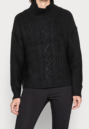 Person, die einen schwarzen Strickpullover mit Rollkragen und schwarze enganliegende Hosen trägt, steht mit entspannten Armen seitlich an einem weißen Hintergrund.