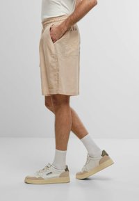 Beige lina šķirtes ar elastīgu jostasvietu, sānu kabatām un diskrētu izšūtu logotipu kreisajā pusē, apvienotas ar baltām sporta kurpēm.