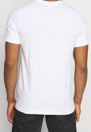Homme vu de dos portant un t-shirt blanc uni à manches courtes et un jean noir, debout sur un fond clair.