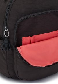 Sac à dos noir avec une poche intérieure corail, une fermeture éclair et un petit porte-clés figurine noire. Tissu texturé avec des détails de couture visibles.