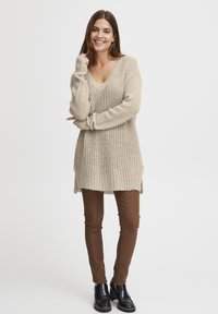 Beżowy sweter dzianinowy z dekoltem w serek i długimi rękawami, zestawiony z brązowymi spodniami w slim fit i czarnymi botkami za kostkę. Teksturowany wzór dzianiny.