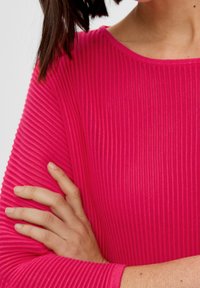 Enganliegendes Langarmoberteil aus glänzendem pinkem Rippstoff, mit einem Bootsausschnitt und strukturiertem Muster. Die Ärmel enden am Handgelenk.