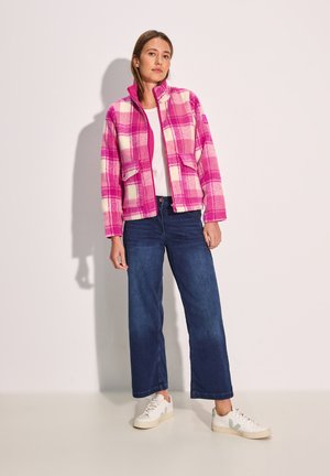 Veste en polaire à carreaux rose et blanc avec col haut, fermeture éclair à l'avant et deux poches, associée à un jean bleu marine à jambes larges et des baskets blanches.