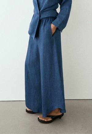 Osoba nosząca luźne, niebieskie spodnie o szerokich nogawkach i dopasowaną górę z ręką w kieszeni, połączone z czarnymi sandałami na obcasie na szarej podłodze.