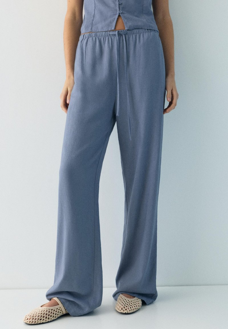 Femme portant un pantalon ample bleu clair à cordon de serrage et des chaussures beiges tissées sans lacets, debout devant un fond clair uni.