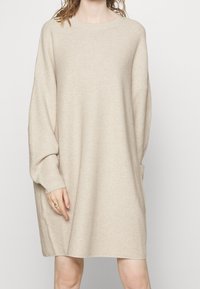 Beige pletené šaty s žebrovanou strukturou, volný střih, dlouhé rukávy a postranní kapsy. Kulatý výstřih a délka do kolen.