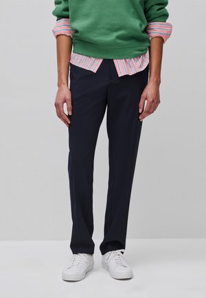 Person trägt schwarze, gerade geschnittene Hose, weiße Sneaker, grünen Pullover über einem rosa gestreiften Hemd, steht vor einfarbigem Hintergrund.