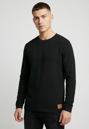 Junger Mann mit kurzen Haaren, der ein schwarzes, strukturiertes Langarmshirt trägt und vor einem schlichten hellgrauen Hintergrund steht.