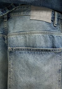 Gros plan sur la poche arrière d’un jean en denim bleu clair avec un écusson en cuir délavé et des coutures apparentes.