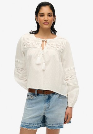 White blouse with textured fabric, lace details along the neckline and sleeves, and drawstring ties. Paired with denim shorts.