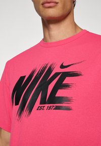 Lyserød bomulds T-shirt med rund halsudskæring, der har et stort sort Nike-logo og "EST. 1972" i fed, penselstrøgs-typografi.
