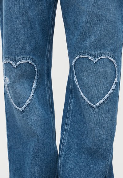 Μπλε τζιν με ξεφτισμένες ανοίγματα σε σχήμα καρδιάς στα γόνατα, που αποκαλύπτουν το ύφασμα από κάτω.