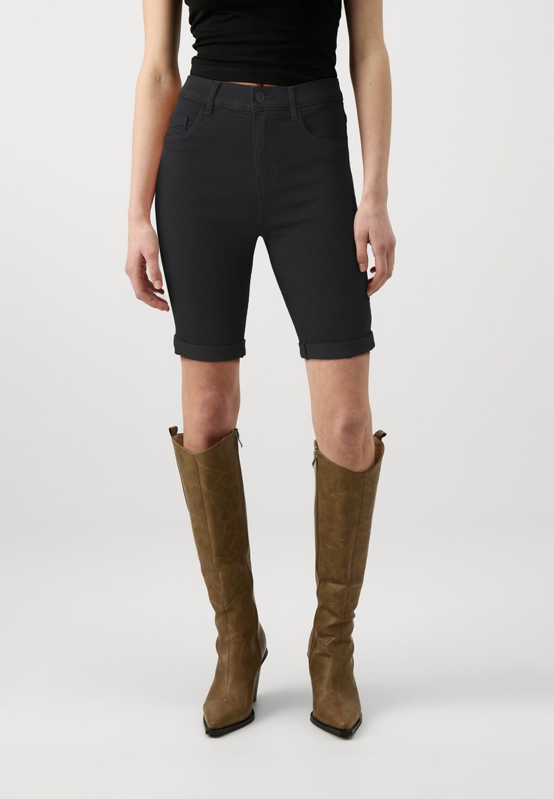 Pantalones cortos de mezclilla negra con puños doblados, combinados con botas marrones hasta la rodilla con punta afilada y costuras decorativas.