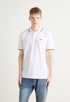 Hvid polo-shirt med sort kant, med en to-knaps knaplukning og broderet logo på venstre bryst. Sammen med blå jeans.