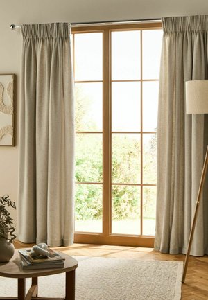 Grande finestra con cornice in legno e tende beige, accanto a una lampada da terra e un tavolo rotondo in legno con libri e decorazioni su un tappeto chiaro.