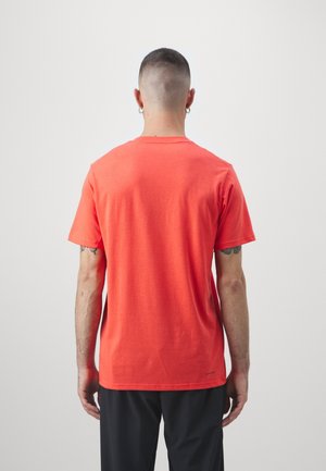 Mężczyzna z krótkimi włosami i kolczykami, ubrany w jednolity czerwony krótki rękaw T-shirt i czarne spodnie, stoi tyłem na białym tle.
