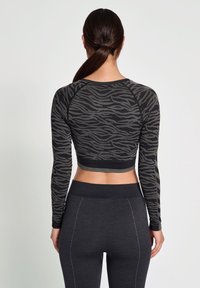 En person står med ryggen mot kameran, iklädd en långärmad, figurnära svart sporttop med ett vågmönster och mörka leggings.