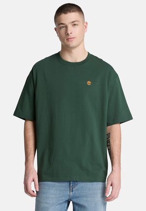 Jonge man met kort haar die een donkergroene oversized T-shirt draagt met een klein oranje logo op de borst en lichtblauwe spijkerbroek.