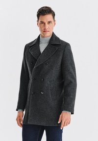 Ciemnoszara wełniana peleryna z podwójnym zapięciem na guziki, szerokimi klapami i dwoma bocznymi kieszeniami. Nosi się ją na jasnoszarym golfie.