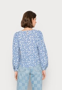 Blusa floral azul com mangas longas, costuras de ombro bufantes e um recorte em forma de chave nas costas. Apresenta pequenas flores brancas sobre um fundo azul claro.