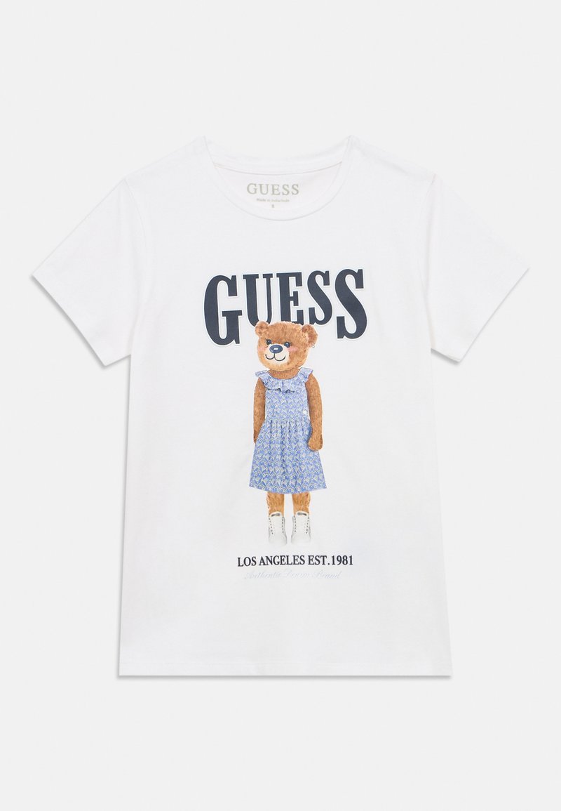 Άσπρο βαμβακερό t-shirt με εικόνα αρκούδας που φοράει ένα μπλε φόρεμα με μοτίβο, με τα λόγια "GUESS" και "LOS ANGELES EST. 1981" από κάτω.