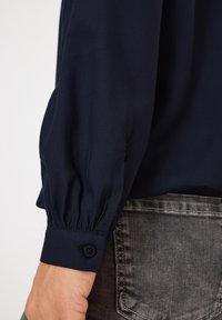 Blouse manches longues bleu marine avec des poignets froncés, dotée d'une fermeture à boutons. Tissu lisse avec un design ajusté et un léger éclat.