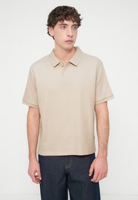 Μπεζ ριμπ πλεκτό πόλο πουκάμισο με γιακά, με δύο κουμπιά στη λαιμόκοψη και κοντά μανίκια. Φορεμένο με σκούρο τζιν τζιν παντελόνι μπροστά σε απλό φόντο.