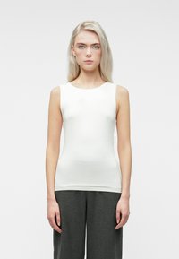 Jeune femme aux longs cheveux blonds, portant un haut sans manches blanc et un pantalon gris foncé, se tenant devant un fond clair uni.