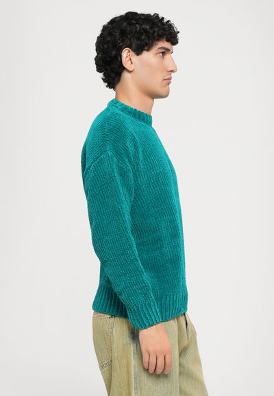 Man with curly hair wearing turquoise sweater and light-colored pants, standing in profile against plain background.
