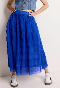 Jupe en tulle bleu à plusieurs couches avec un design plissé, taille élastique et texture transparente. Associée à des baskets blanches avec des rayures noires.