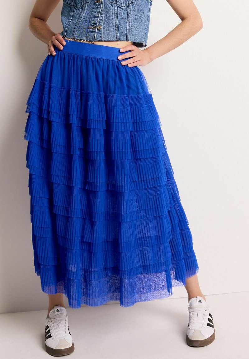 Jupe en tulle bleu à plusieurs couches avec un design plissé, taille élastique et texture transparente. Associée à des baskets blanches avec des rayures noires.