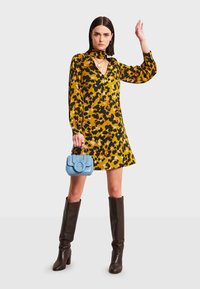 Nainen pukeutuneena keltaiseen ja mustaan kukalliseen pitkähiharaiseen mekkoon, polven ylle ulottuviin ruskeisiin saappaisiin, pitäen pientä sinistä käsilaukkua, toinen käsi nostettuna.