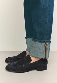 Mocassini neri in camoscio con tacco piatto, caratterizzati da un dettaglio cucito sulla parte superiore. Indossati con pantaloni in denim scuro, risvoltati con tessuto a contrasto.
