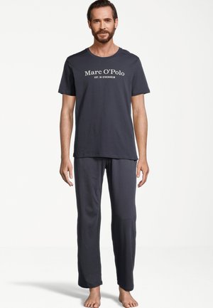 Mężczyzna z brodą, ubrany w ciemnoszarą koszulkę Marc O’Polo i pasujące luźne spodnie, stojący boso na jednolitym białym tle.