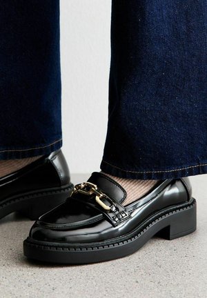 Black patent leather loafers with a chunky heel, featuring a gold chain accent. The shoe has a rounded toe and a textured sole.