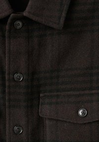 Camisa de franela marrón con patrón de cuadros oscuros, que cuenta con cuello, cuatro botones negros y un bolsillo en el pecho. Textura suave.