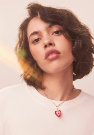 Junge Frau mit kurzen lockigen Haaren, die ein weißes Oberteil und eine goldene Kette mit einem roten Herzanhänger trägt. Weiches, warmes Licht.