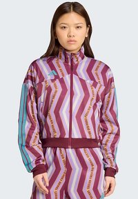 Νεαρή γυναίκα φοράει ένα μωβ και λεβάντα φούτερ με ζιγκ-ζαγκ σχέδιο και ταιριαστά παντελόνια με τυρκουάζ ρίγες στα μανίκια, που στέκεται σε έναν απλό φόντο.