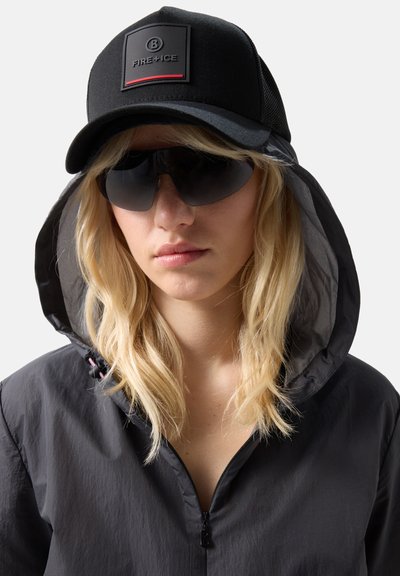 Kvinde med blondt hår iført sort hættejakke, sort kasket med "FIRE+ICE" logo og store sorte solbriller mod en ensfarvet baggrund.