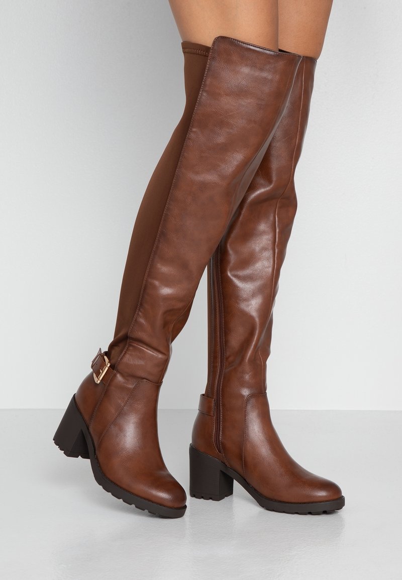 Botas marrones de cuero por encima de la rodilla con tacón grueso, cremallera lateral y un adorno de hebilla ajustable en el tobillo. Textura lisa y diseño ajustado.