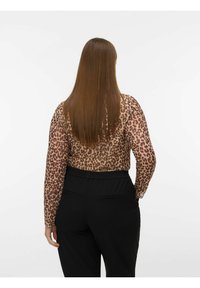 Vrouw staat met haar rug naar de camera, draagt een lange mouwen top met luipaardprint en zwarte broek, met lang, steil bruin haar.