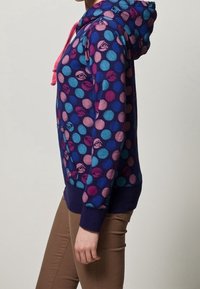 Sweat à capuche violet avec des pois colorés en rose, bleu et violet. Dispose d'une capuche à cordon de serrage et de poignets côtelés. Texture de tissu douce.