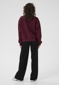 Felpa bordeaux con vestibilità rilassata e polsini a costine, abbinata a pantaloni neri a gamba larga. Texture liscia, dettagli minimi e design semplice.
