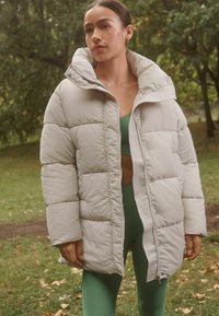 Lys grå puffjakke med høj krave, lynlås foran og quiltet design. Bæres over en grøn top og leggings med en afslappet pasform.