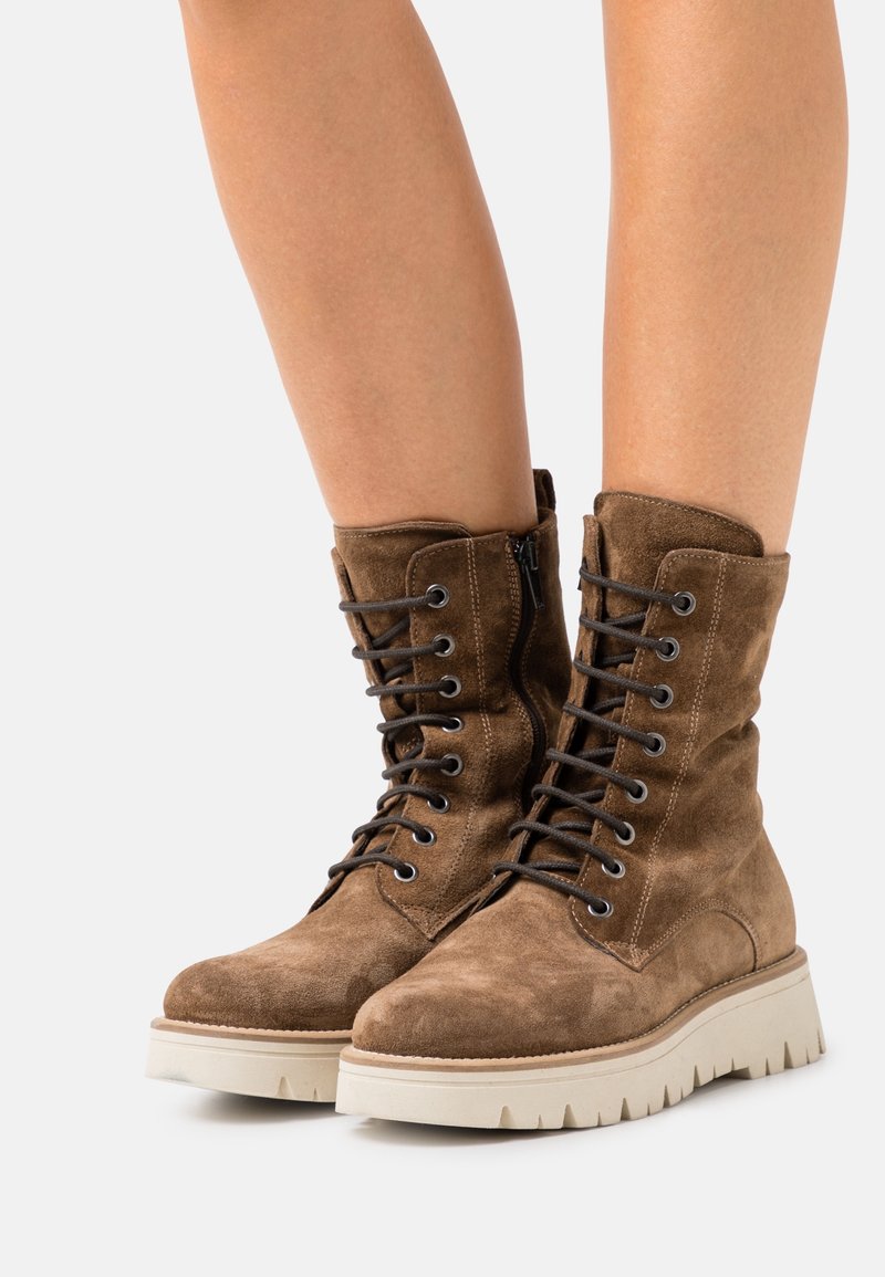 Bottes cheville en daim marron à lacets avec fermeture éclair latérale et semelles épaisses blanches à rainures, portées sur jambes nues.