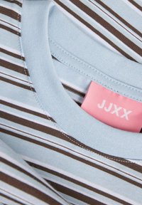 Tricou bleu deschis cu guler rotund, cu dungi orizontale maro și albe și o etichetă roz în interiorul gulerului.